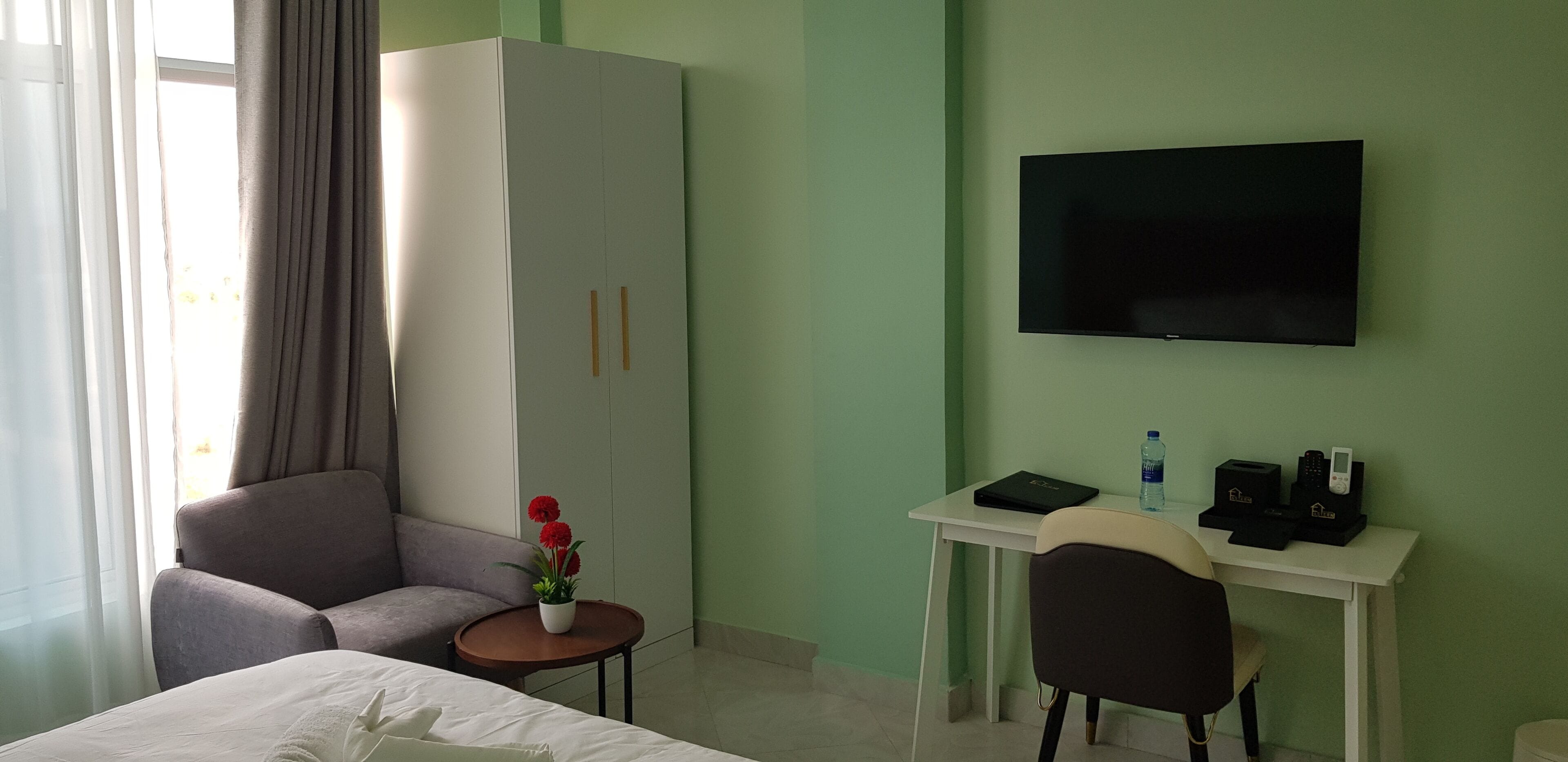 Ett hotellrum med en säng, en soffa, ett skrivbord med en tv, ett litet bord med en växt och en garderob.