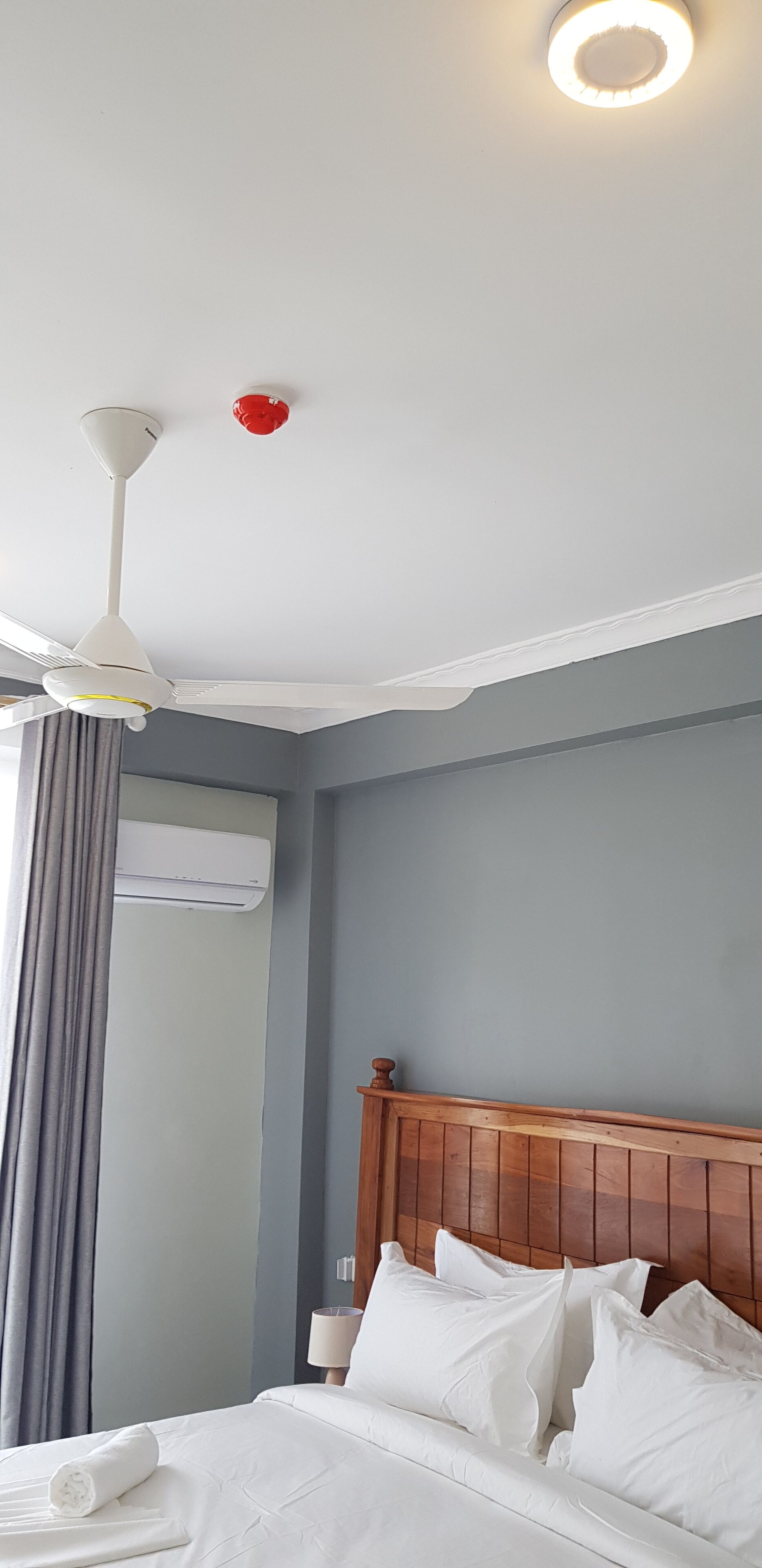 Una habitación de hotel con cama, cabecero de madera, ventilador de techo, equipo de aire acondicionado y ventana con cortinas.