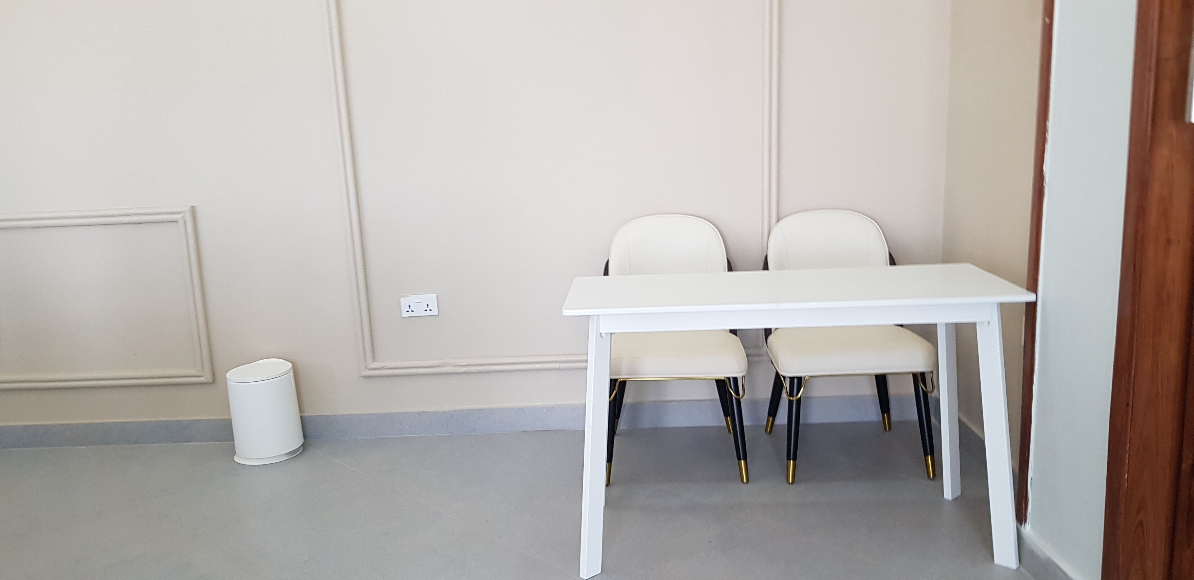 A white table with two chairs in a corner of a room with beige walls and a grey floor.
