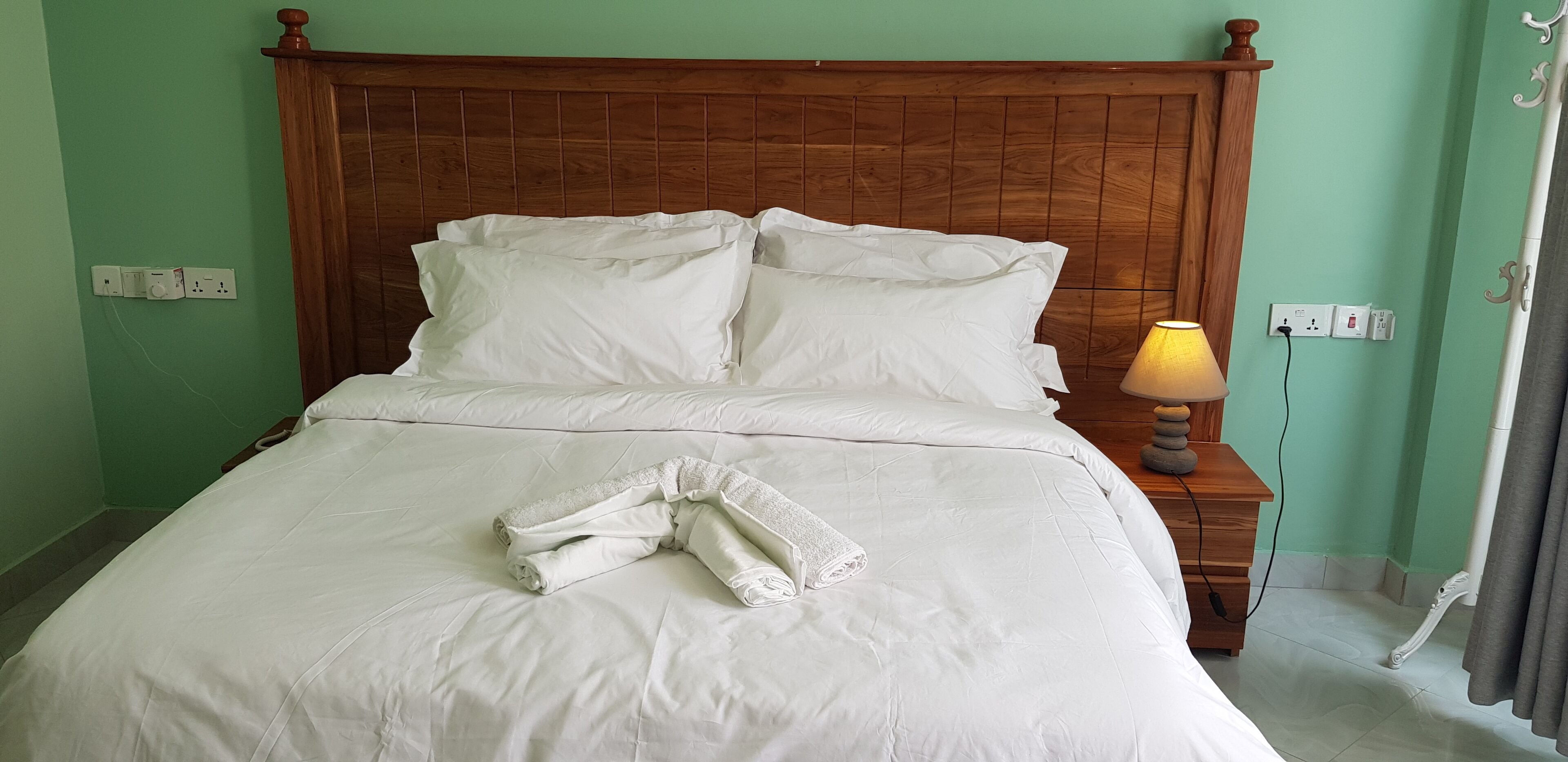 A neatly made bed with white linens and a rolled towel, a wooden headboard, and a bedside lamp.