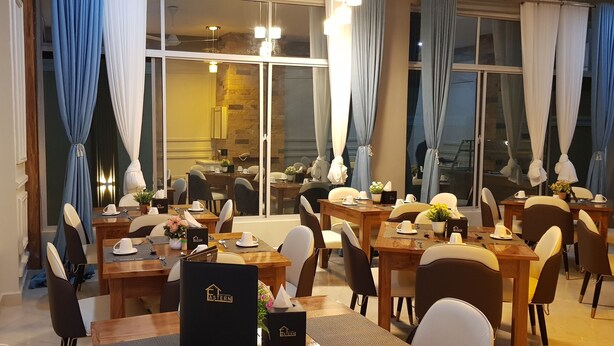 Comedor de hotel con varias mesas dispuestas para los huéspedes, un televisor montado en la pared y grandes ventanales con cortinas.