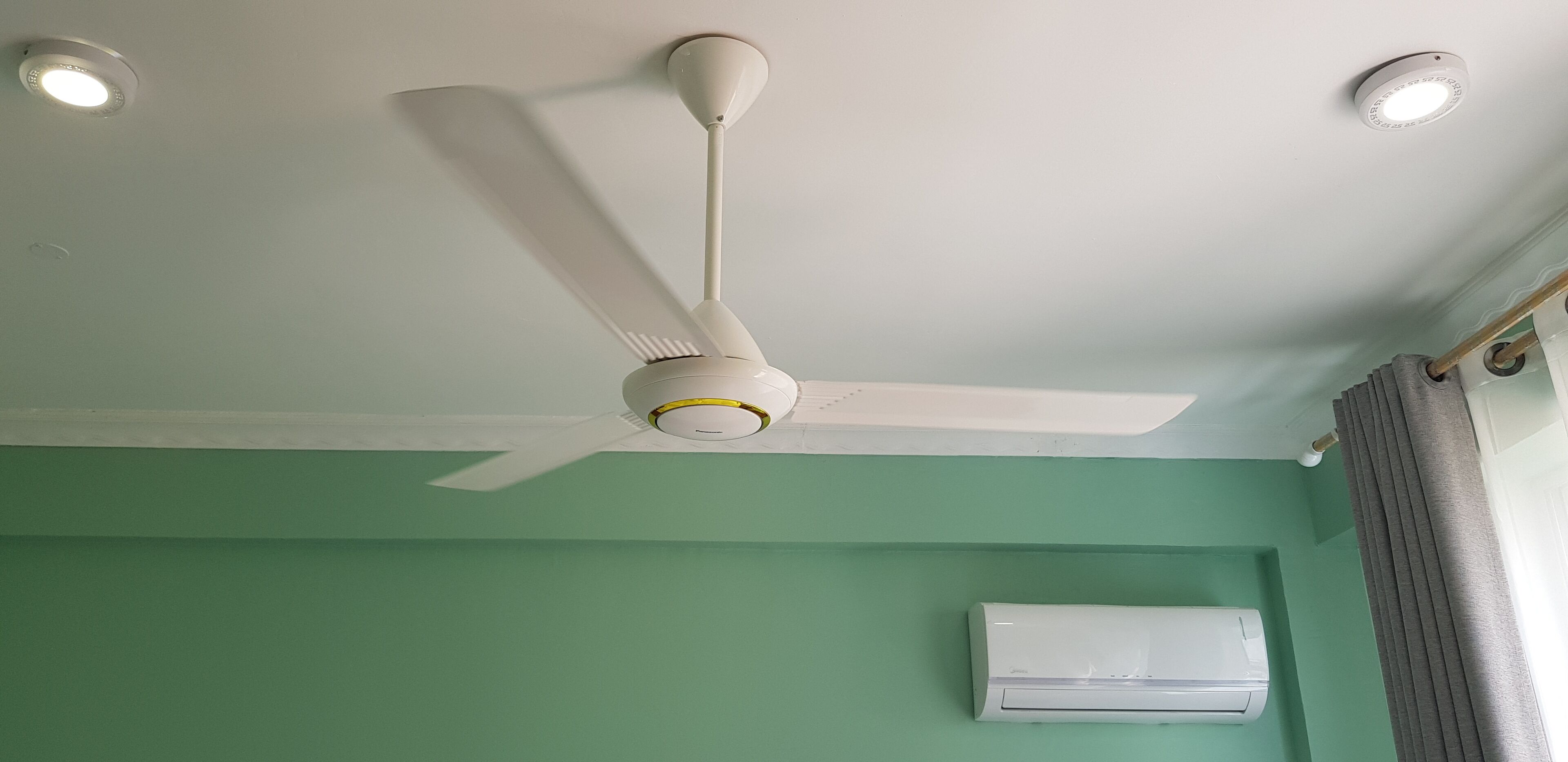 Ein Deckenventilator mit drei Lampen, eine weiße Klimaanlage und eine grüne Wand.