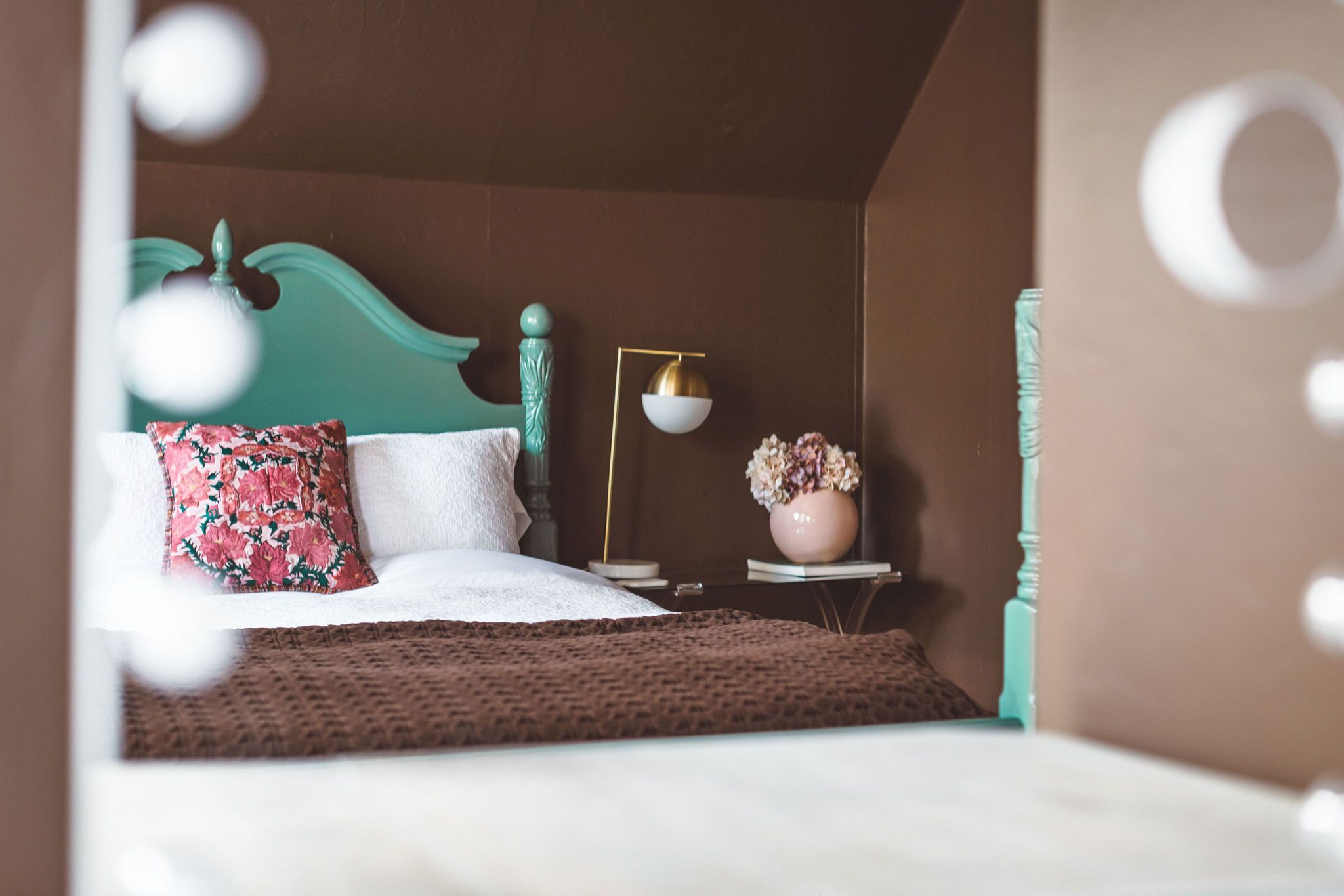 A neatly made bed with a floral pillow, a bedside lamp, and a vase with flowers on a small table beside the bed.