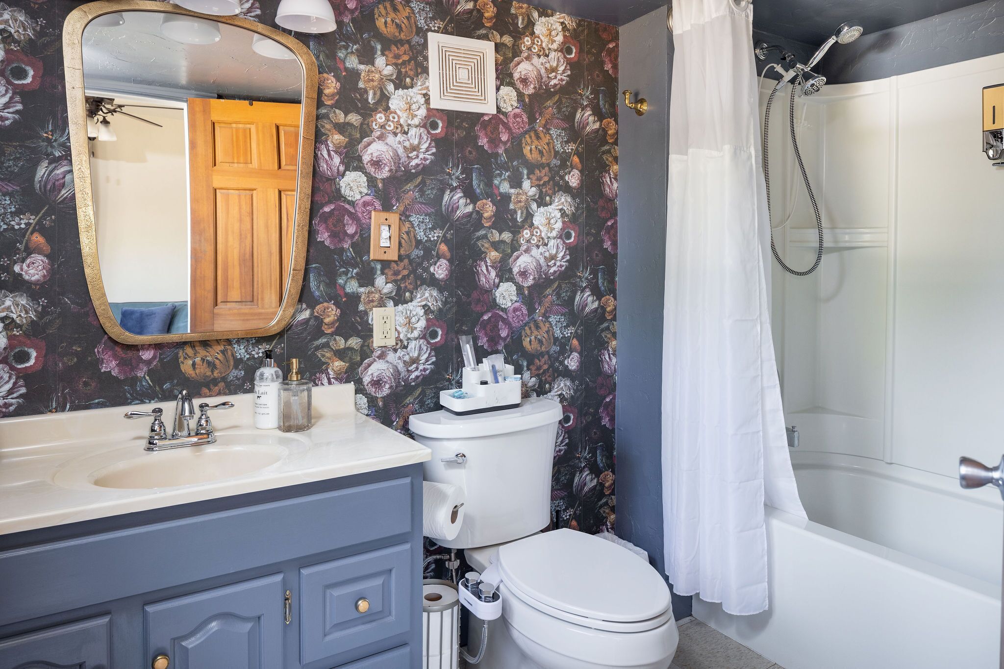 Une salle de bain avec un papier peint à motifs floraux, un lavabo avec un miroir, des toilettes et une baignoire équipée d’un pommeau de douche.