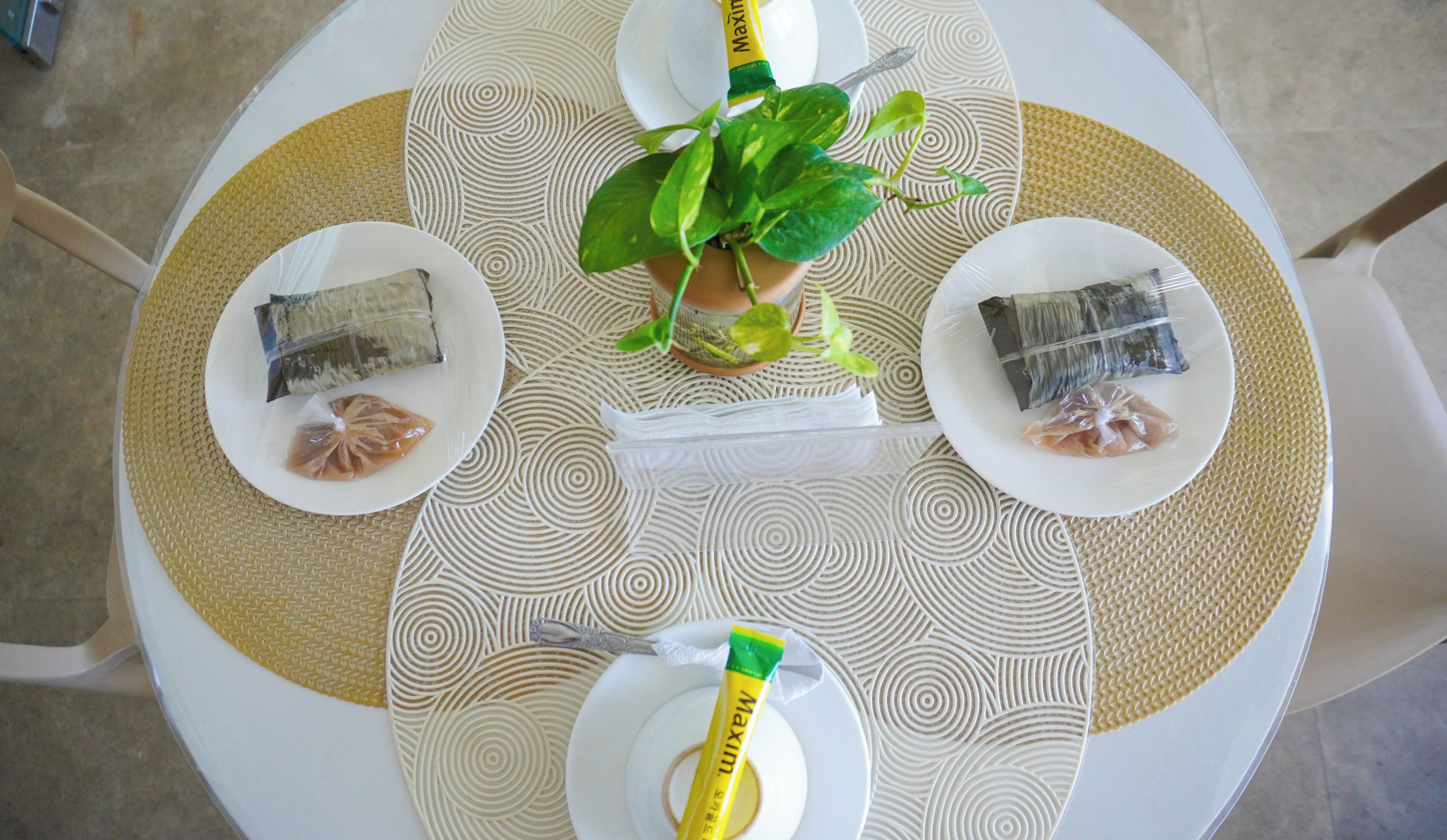 A round table with a white and beige patterned tablecloth, two plates with wrapped food items, a green plant in a pot, and a yellow tube on the table.