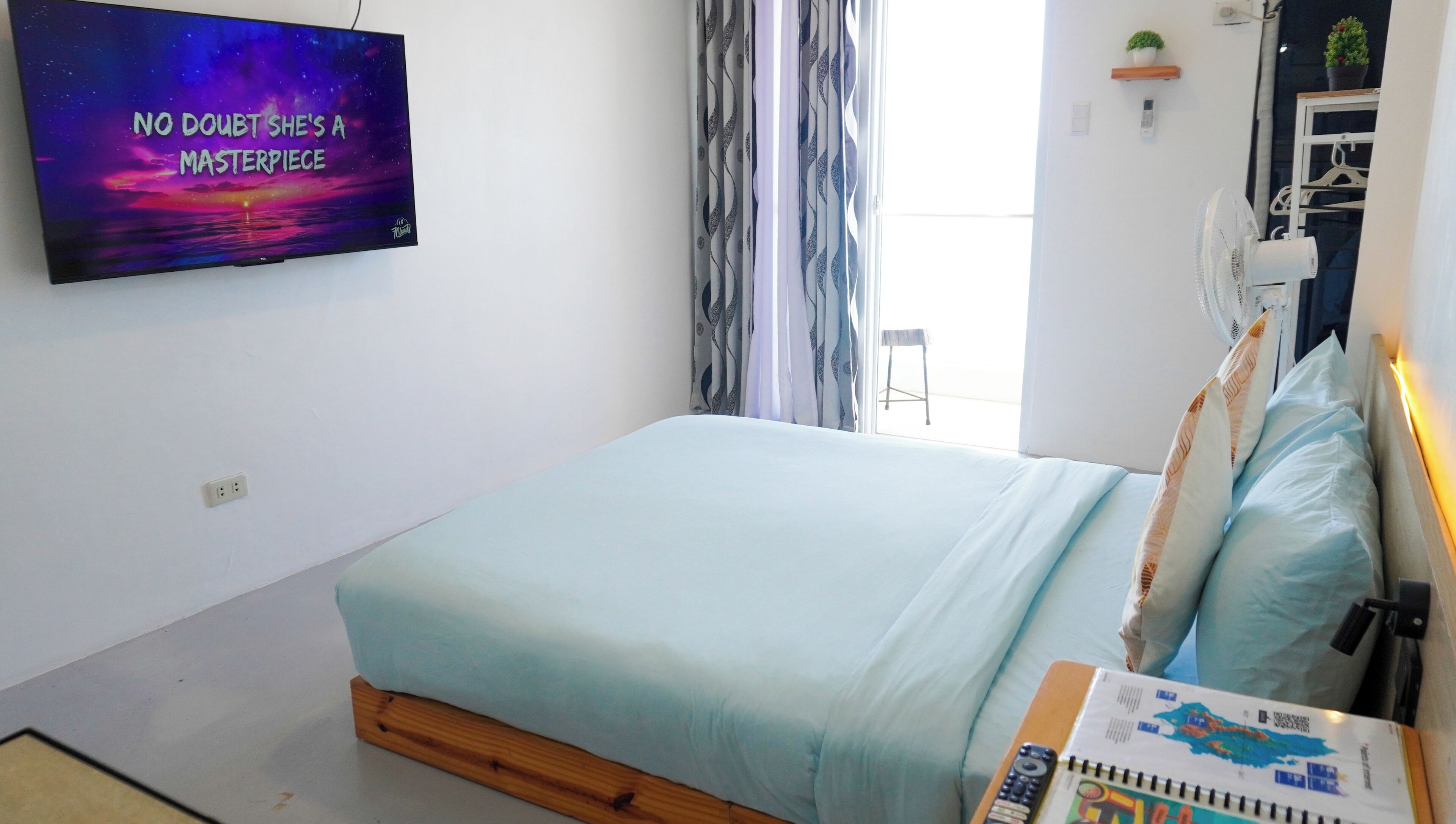 A bedroom with a bed, a TV on the wall, a window with curtains, and a small plant on a shelf.