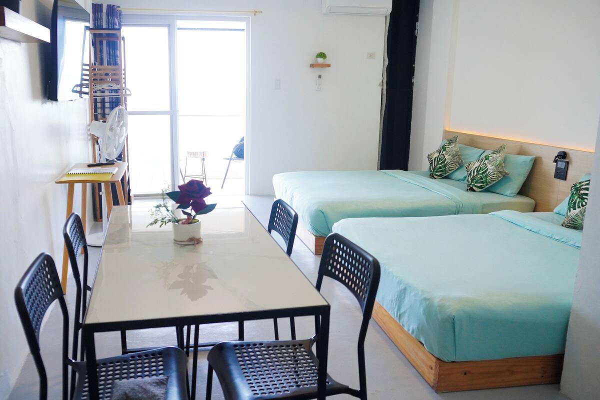 A room with a dining table, two beds, a fan, and a window.