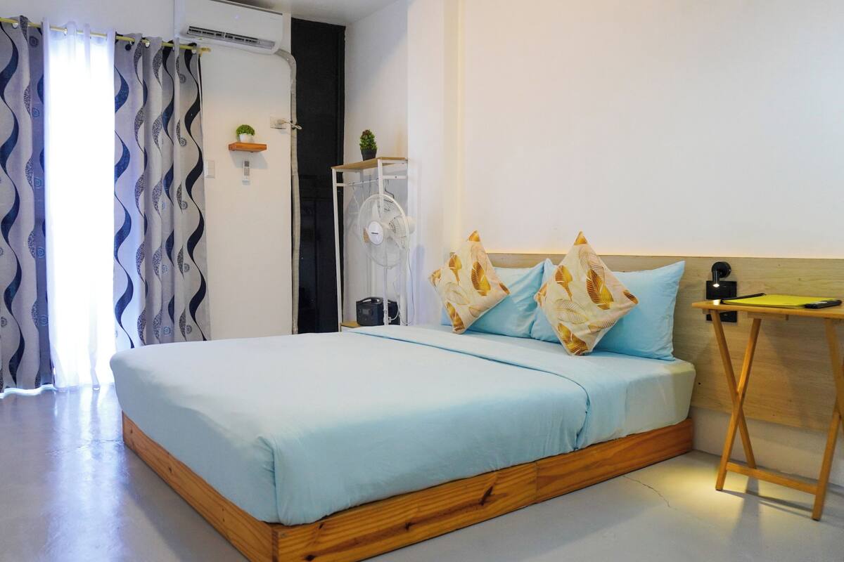 A bedroom with a bed, a bedside table, a fan, and a small shelf with plants.