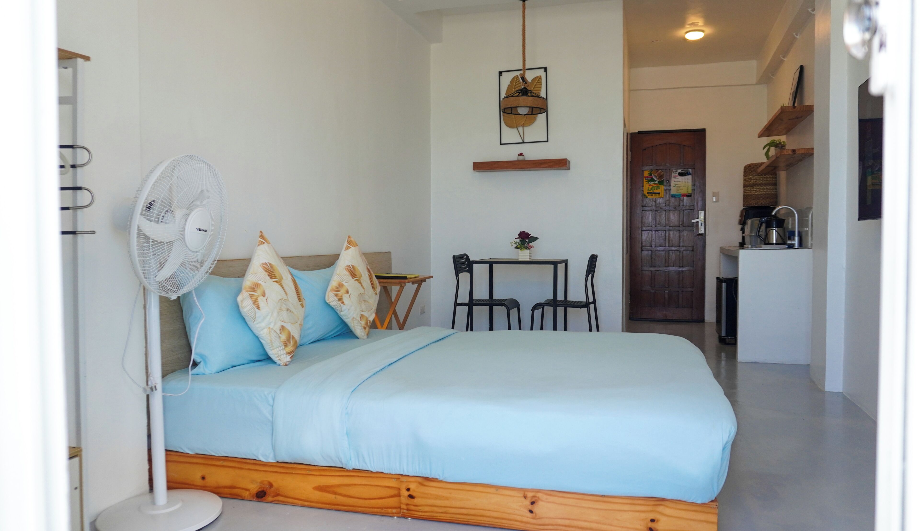 Une chambre avec un lit, un ventilateur, une table avec des chaises, et un coin cuisine à l’arrière-plan.