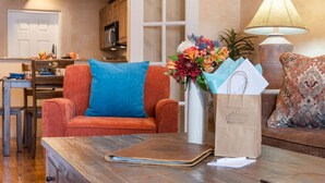 Un salon chaleureux avec une table basse en bois, un fauteuil rouge agrémenté d’un coussin bleu, un canapé avec des coussins à motifs, un vase rempli de fleurs et une lampe.