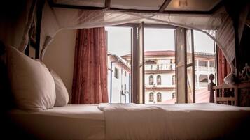 A bed with a canopy, a view of buildings outside, and a window with curtains.