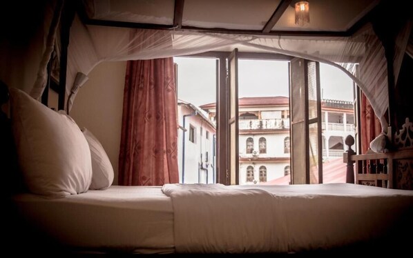 A bed with a canopy, a view of buildings outside, and a window with curtains.