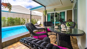 A poolside area with wicker chairs and a glass table, a hanging plant, and a view of a building in the background. - New 2 Bedroom Pool Villa C882 (Hua Hin)