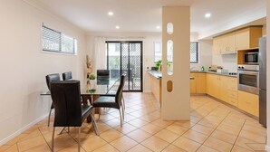 A modern kitchen with a dining area, featuring a glass table, black chairs, and a view through a sliding door to an outdoor balcony. - Happy haven at Eight Mile (Eight Mile Plains)