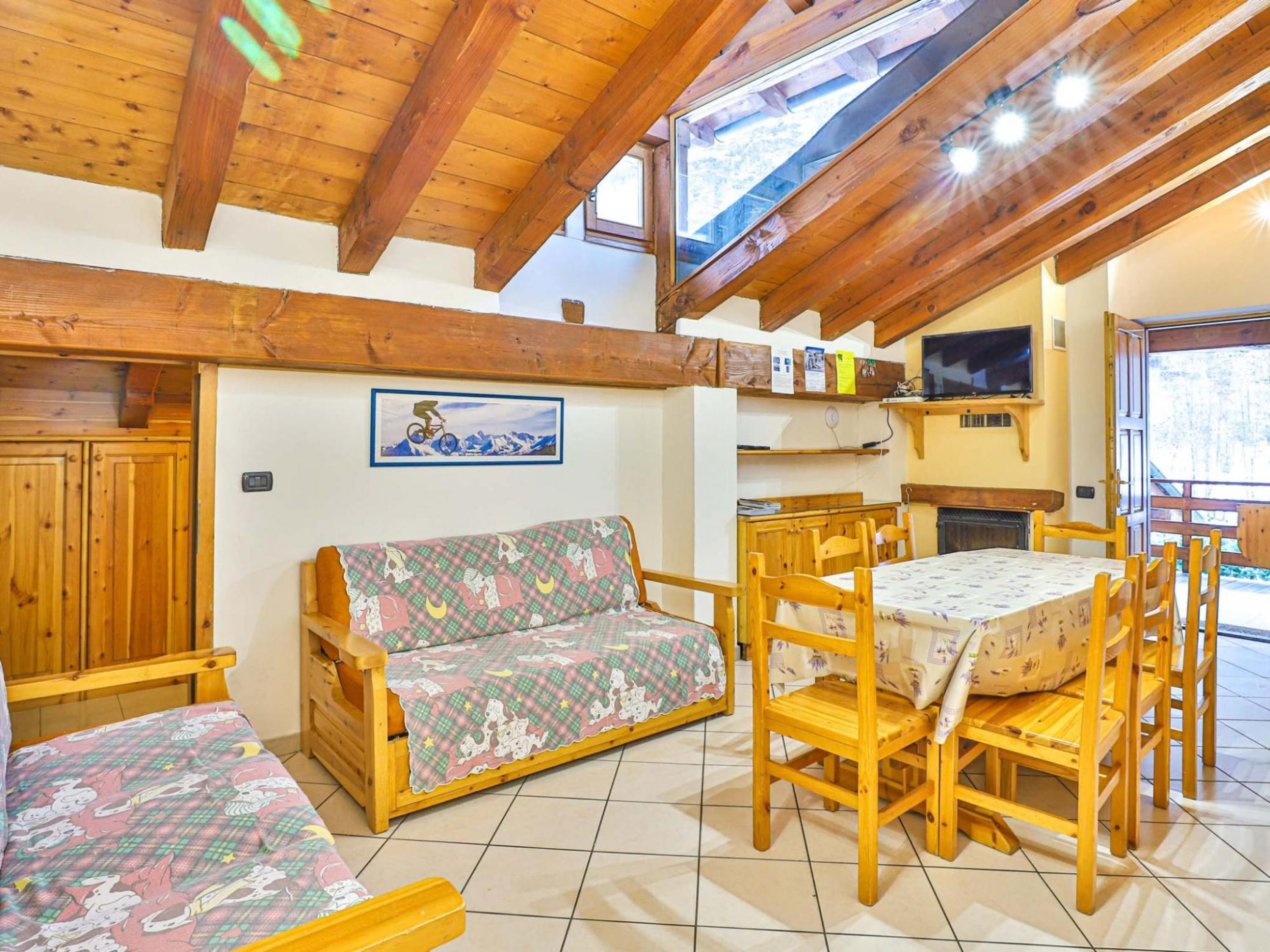 A cozy room with a wooden ceiling, a sofa, a dining table with chairs, a television, and a window.