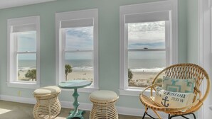 A room with three large windows overlooking a beach, wicker furniture, and a small table. - 109 Grand Pavilion by Avantstay Beachfront Views (Isle of Palms)