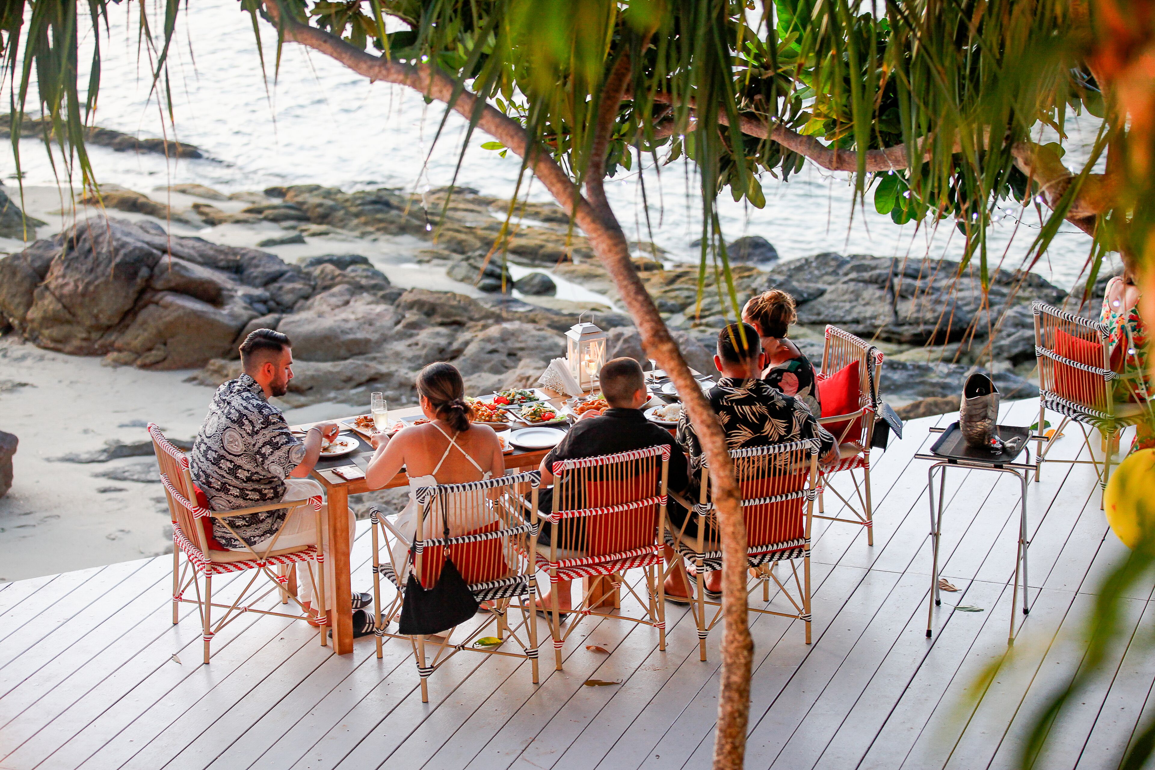 Un grupo de personas cenando en una terraza de madera con sillas de mimbre, con vista a una playa rocosa.