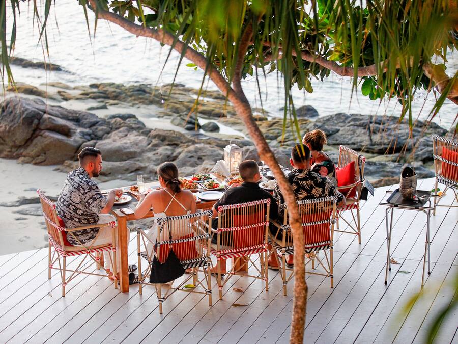Um grupo de pessoas jantando em um deck de madeira com cadeiras de vime, com vista para uma praia rochosa.