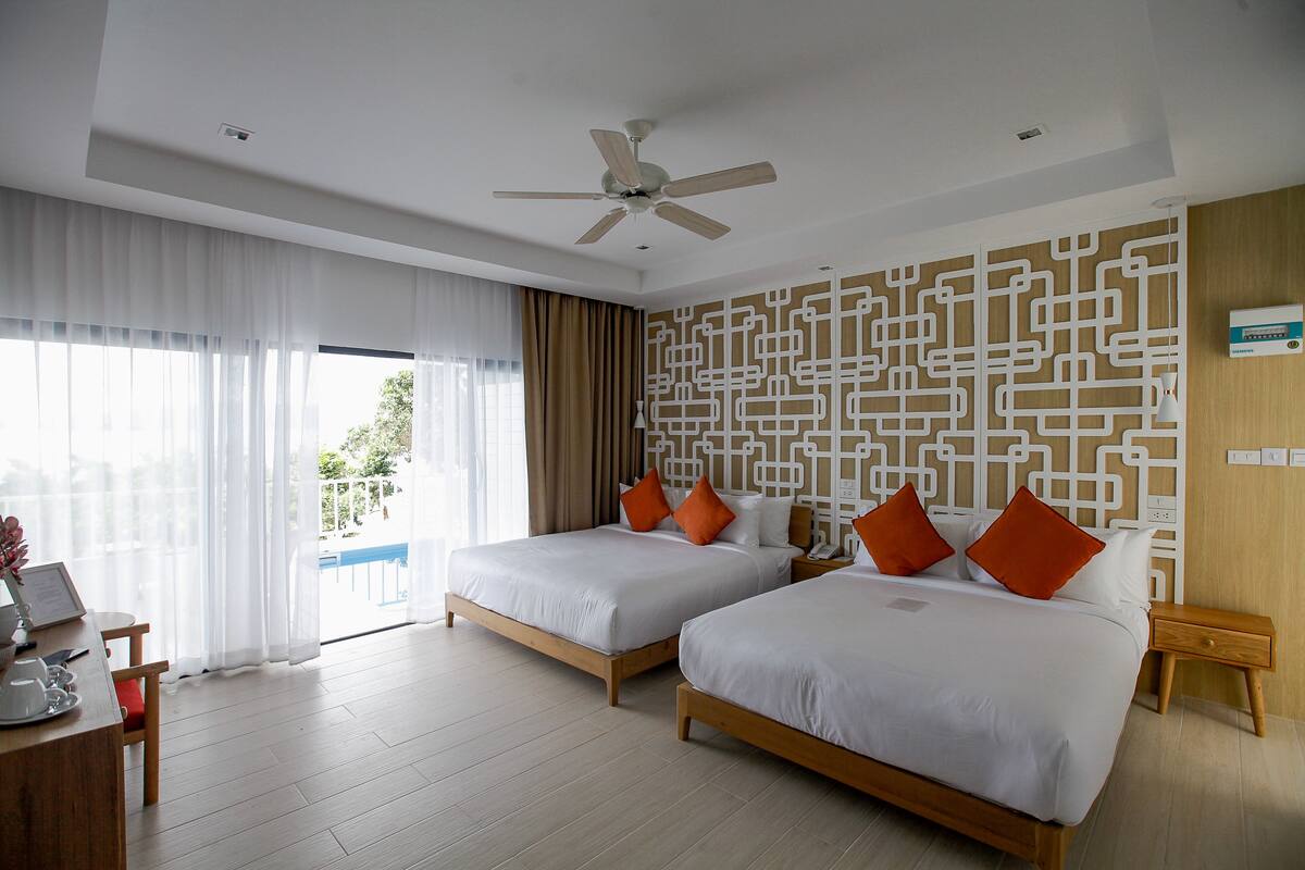 A hotel room with two beds, a desk with a chair, a ceiling fan, and a view of the outdoors.