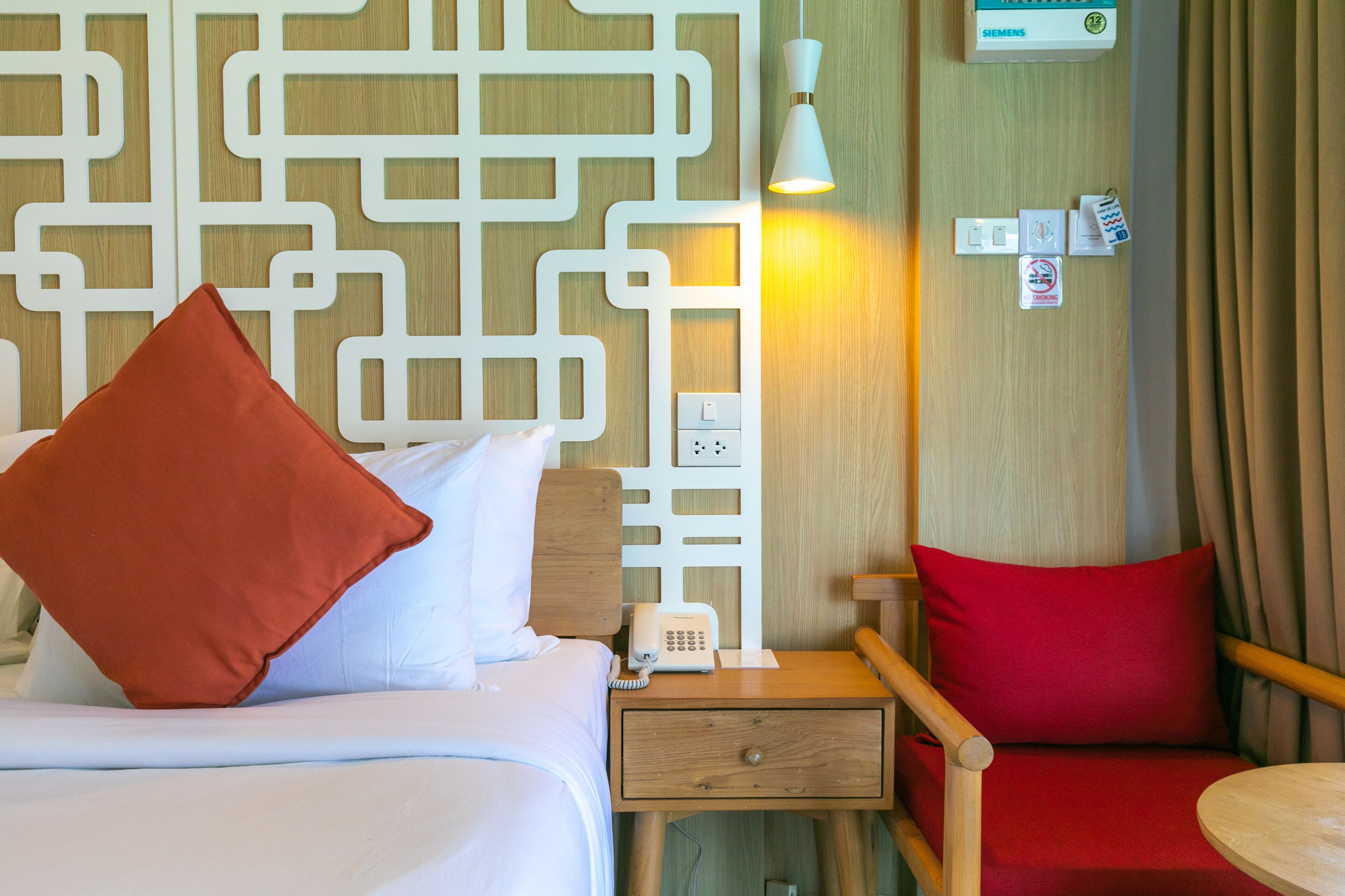 A hotel room with a bed, a nightstand with a phone, a red chair, a small table, and a wall-mounted light fixture.