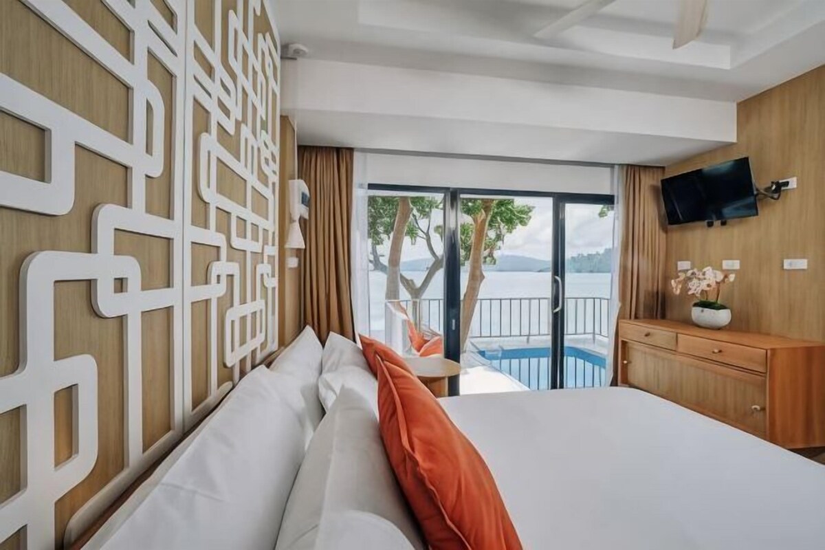 A hotel room with a bed, a TV, a wooden wall panel, a view of the outdoors, and a pool.