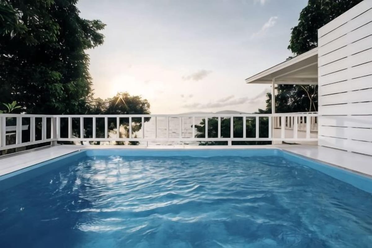 A swimming pool with a white deck and railing, surrounded by greenery and a white building.