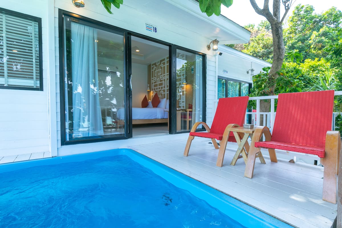 A poolside area with a clear view of a bedroom through glass doors, two red wicker chairs, and a wooden table.