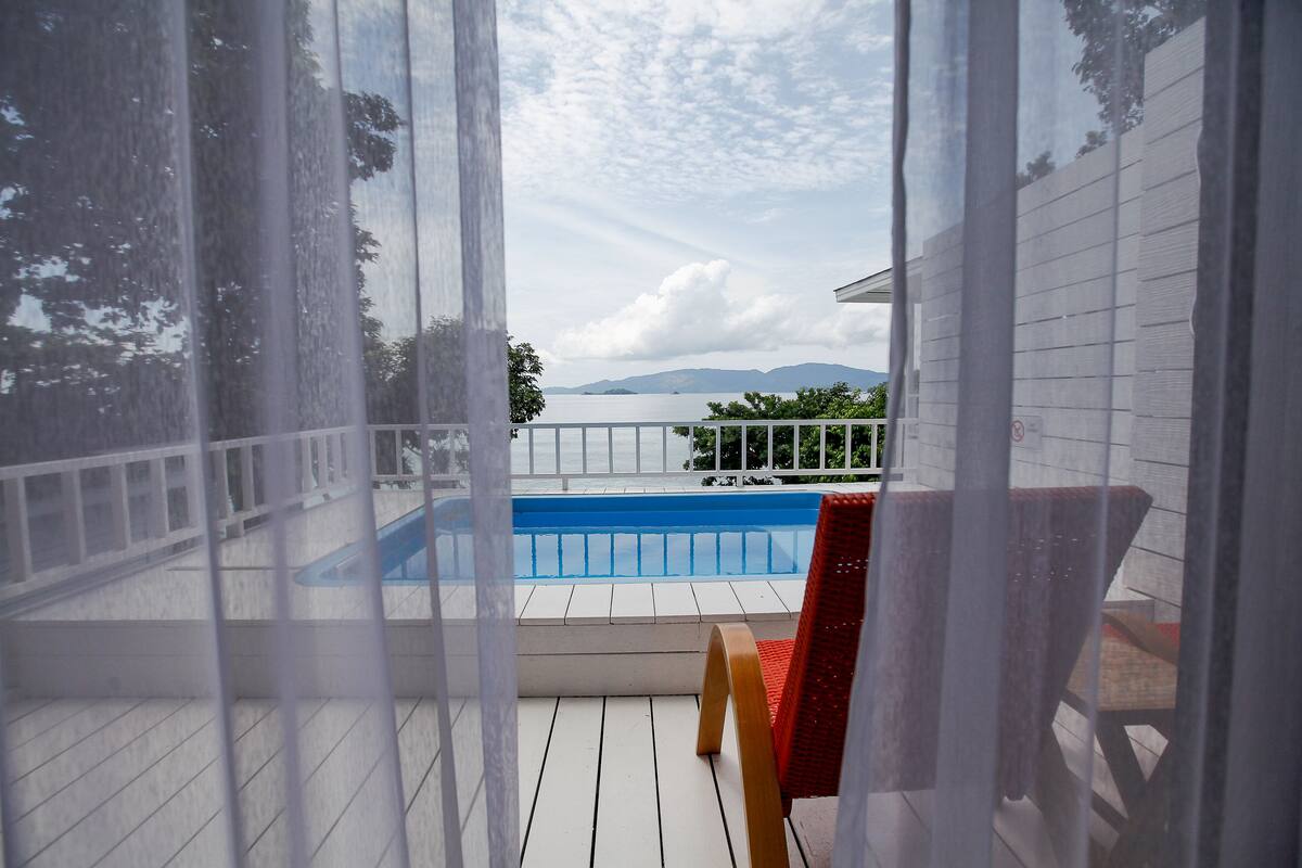 A view of a swimming pool from a covered deck area with a red chair and white railing.