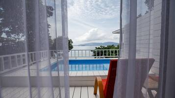 A view of a swimming pool from a covered deck area with a red chair and white railing.