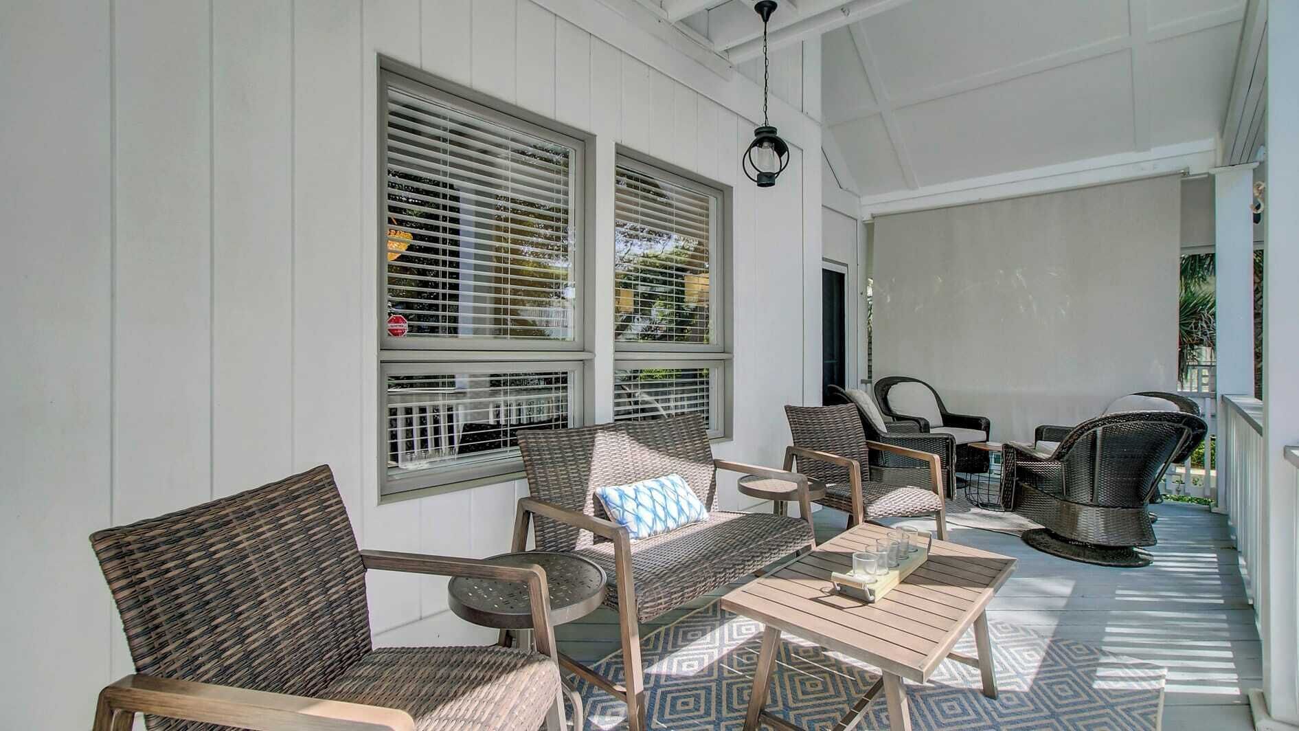 A covered porch with wicker furniture, a table with glasses, and a view of the outdoors through large windows with blinds.