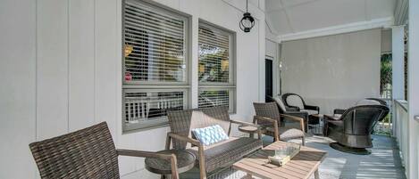 A covered porch with wicker furniture, a table with glasses, and a view of the outdoors through large windows with blinds.