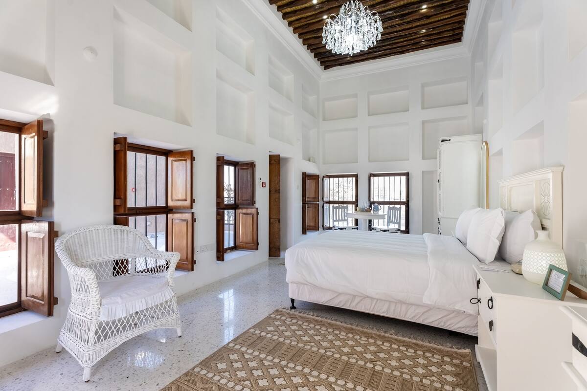 Un dormitorio espacioso con una cama grande, una silla de mimbre blanca y un candelabro.