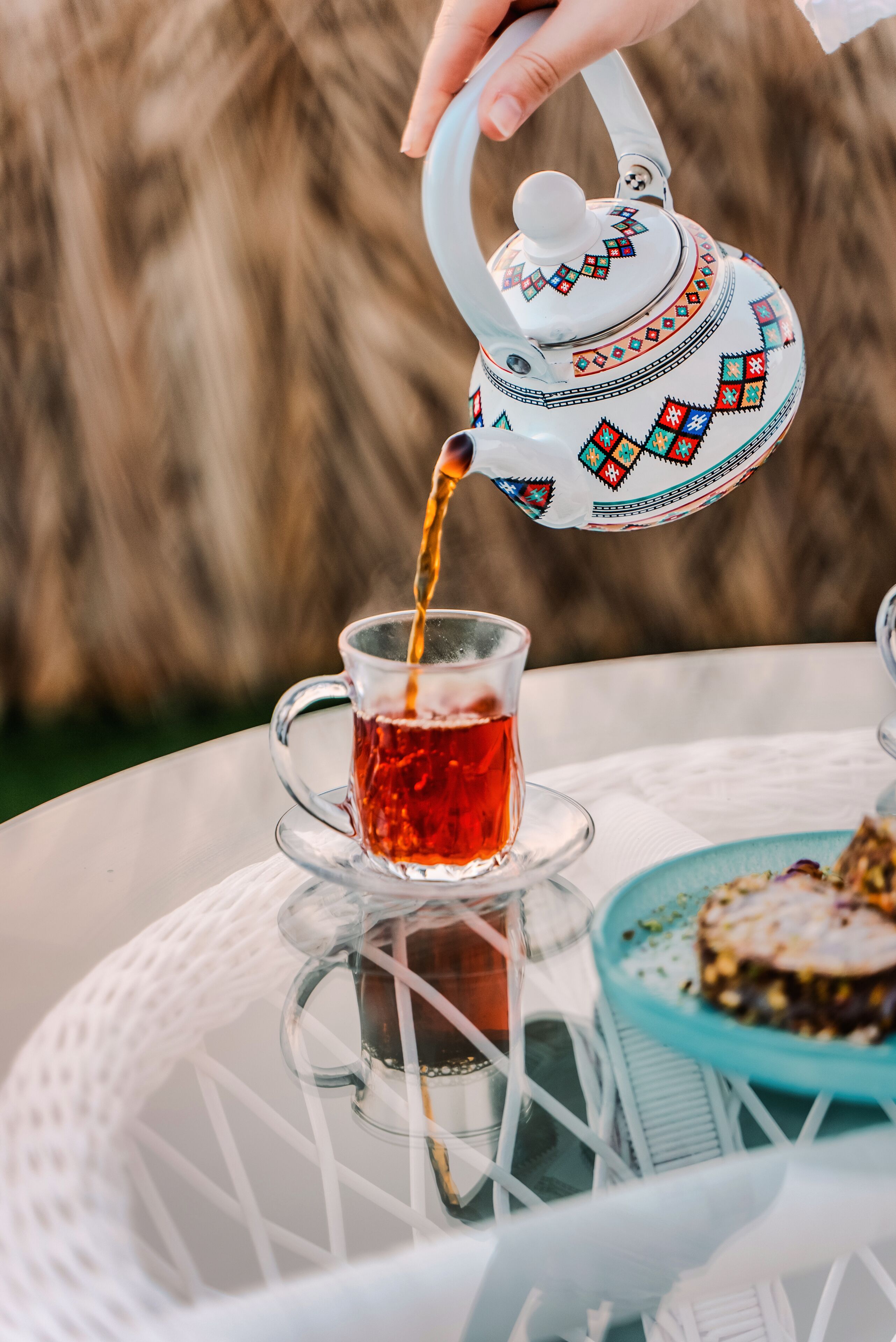A hand pouring tea from a patterned teapot into a clear glass cup on a white table.