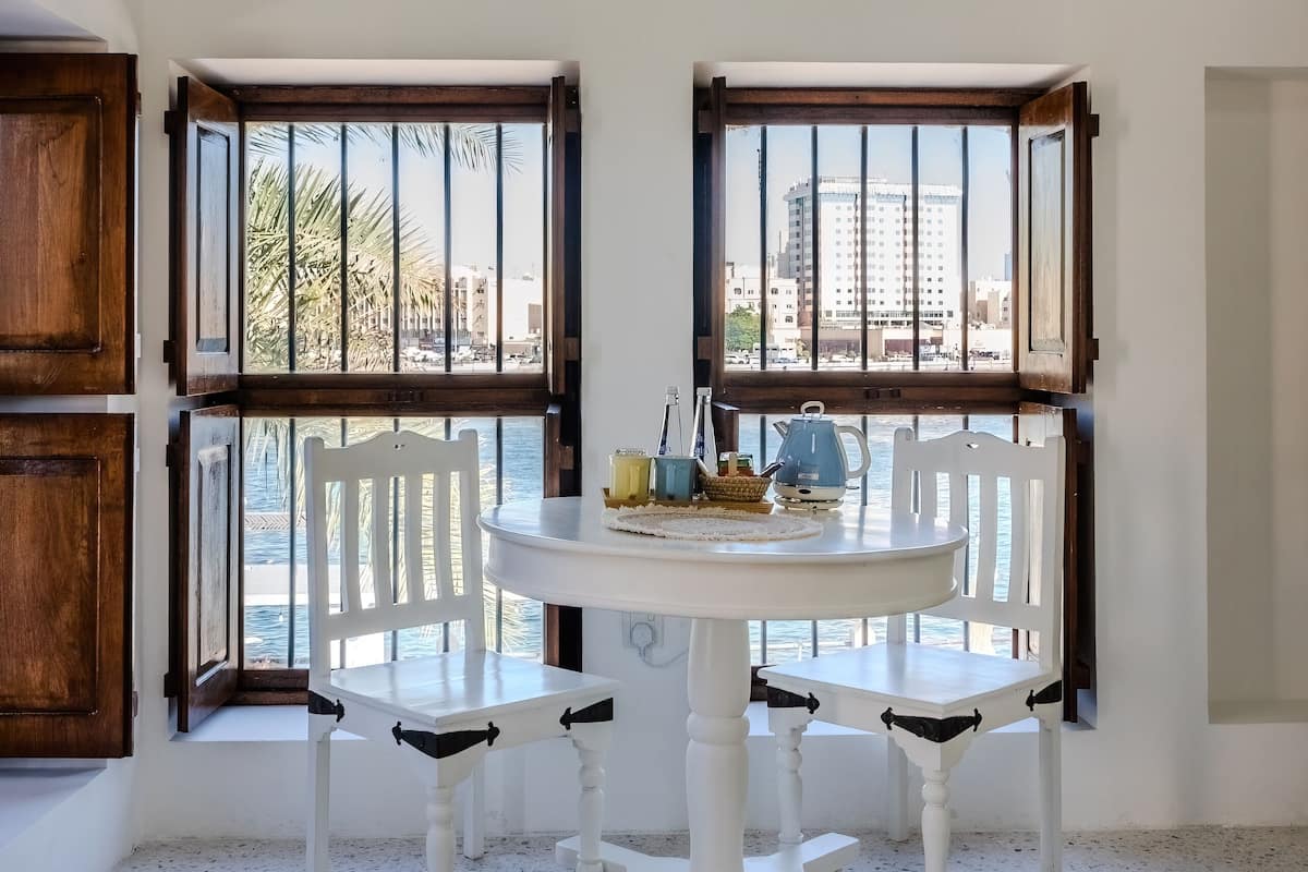 Una habitación con una mesa y sillas blancas, ventanas amplias con marcos de madera y vista a palmeras y edificios.