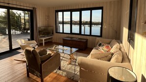 A spacious living room with a large window offering a view of a body of water, wooden flooring, a sofa, a coffee table, and a television. - Beautiful wooden house with sea view (Gujan-Mestras)