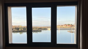 A room with a large window overlooking a calm body of water and distant buildings. - Beautiful wooden house with sea view (Gujan-Mestras)