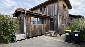 A rustic wooden building with a sloped roof, a front entrance with steps, and two wheelie bins outside. - Beautiful wooden house with sea view (Gujan-Mestras)