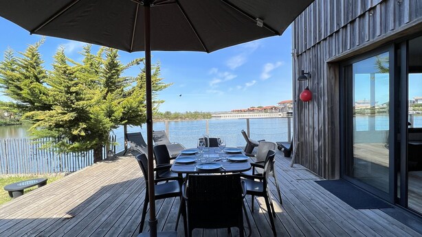 A wooden deck with a table set for dining, overlooking a body of water and a distant shoreline. - Beautiful wooden house with sea view (Gujan-Mestras)