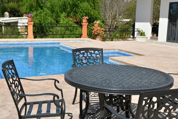Un espace aménagé au bord de la piscine, avec une table ronde et des chaises, entouré d’une clôture et agrémenté de plantes en pot.
