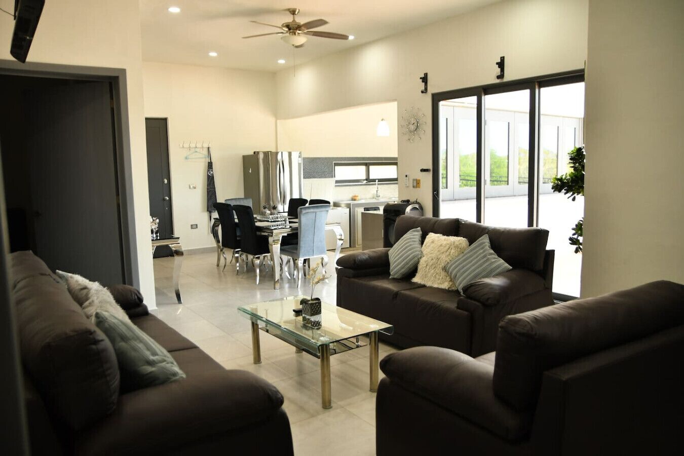 A spacious living area with a dark brown leather sofa set, a glass coffee table, and a dining area with a modern kitchen in the background.