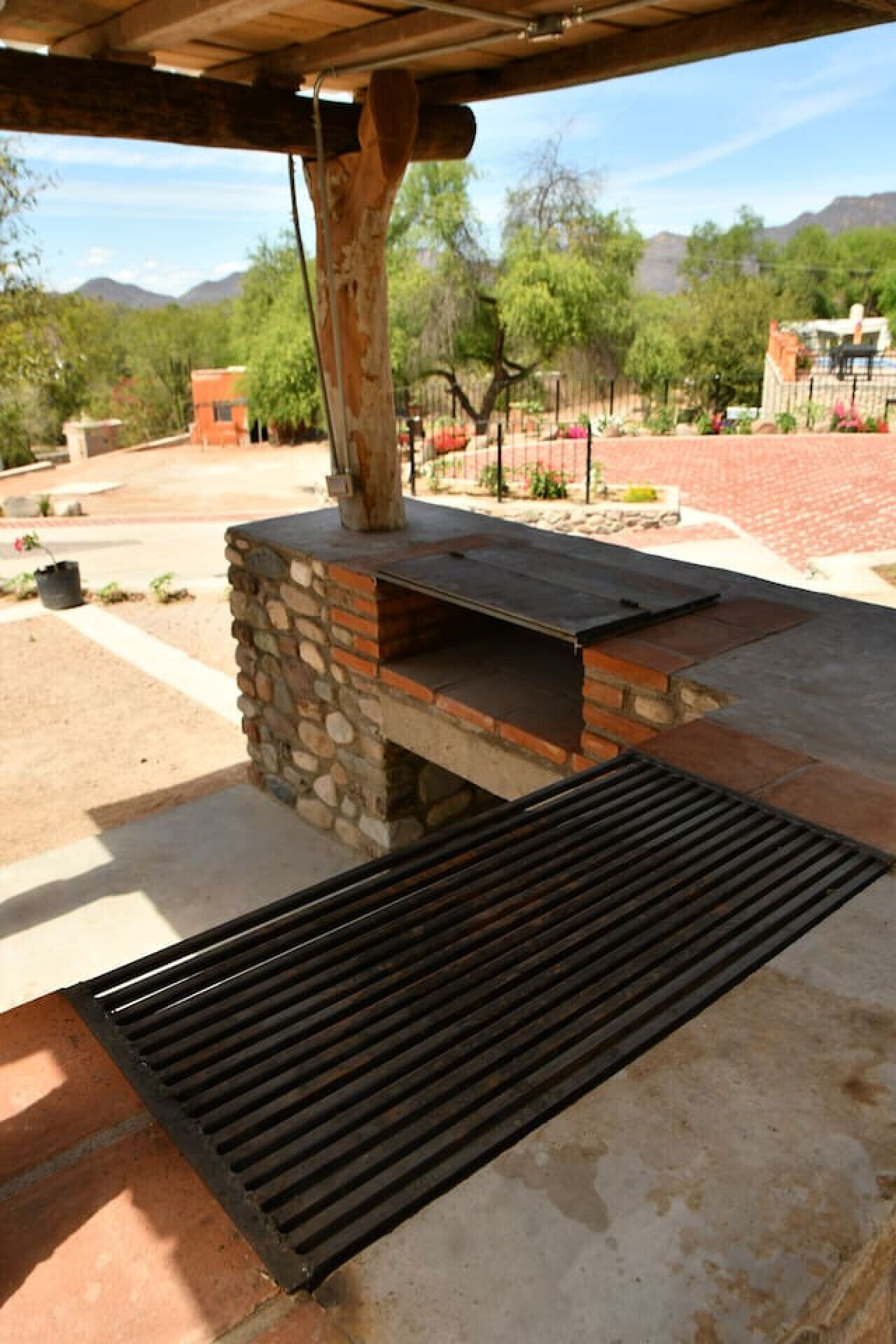 An outdoor barbecue area with a stone and brick structure, a metal grill, and a view of a landscaped garden and mountains in the background.