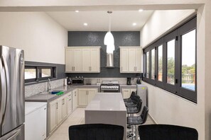 A modern kitchen with a central island, white cabinets, a stainless steel refrigerator, and a large window providing a view of the outdoors.