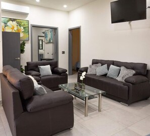 A modern living room with two dark brown sofas, a glass coffee table, and a flat-screen TV mounted on the wall.