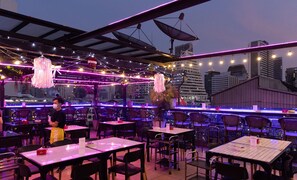 An outdoor dining area with tables and chairs, string lights, and a city view in the background. - Heaven Boutique Hotel (Bangkok)