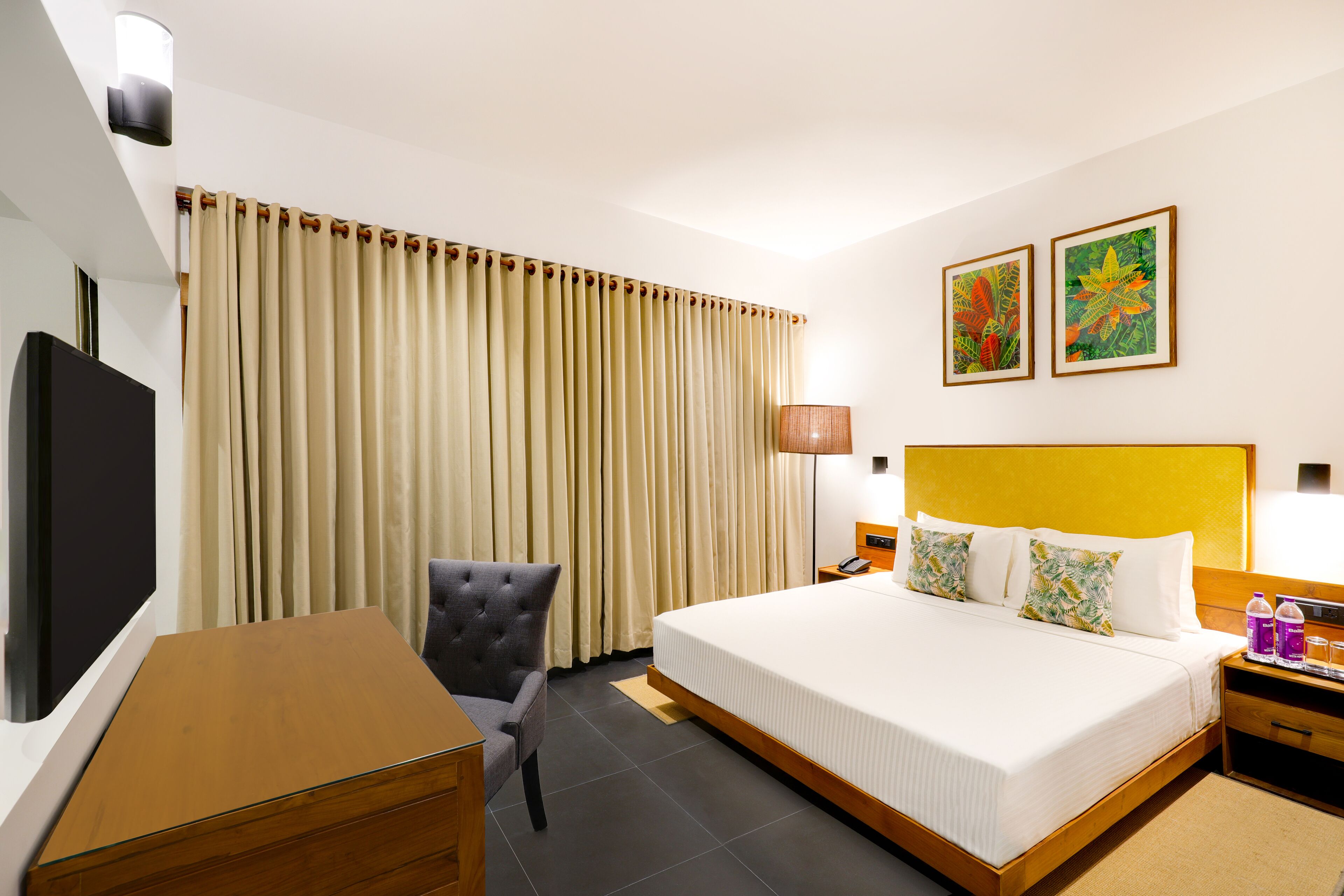A hotel room with a bed, a desk, a chair, a TV, a lamp, and two paintings on the wall.