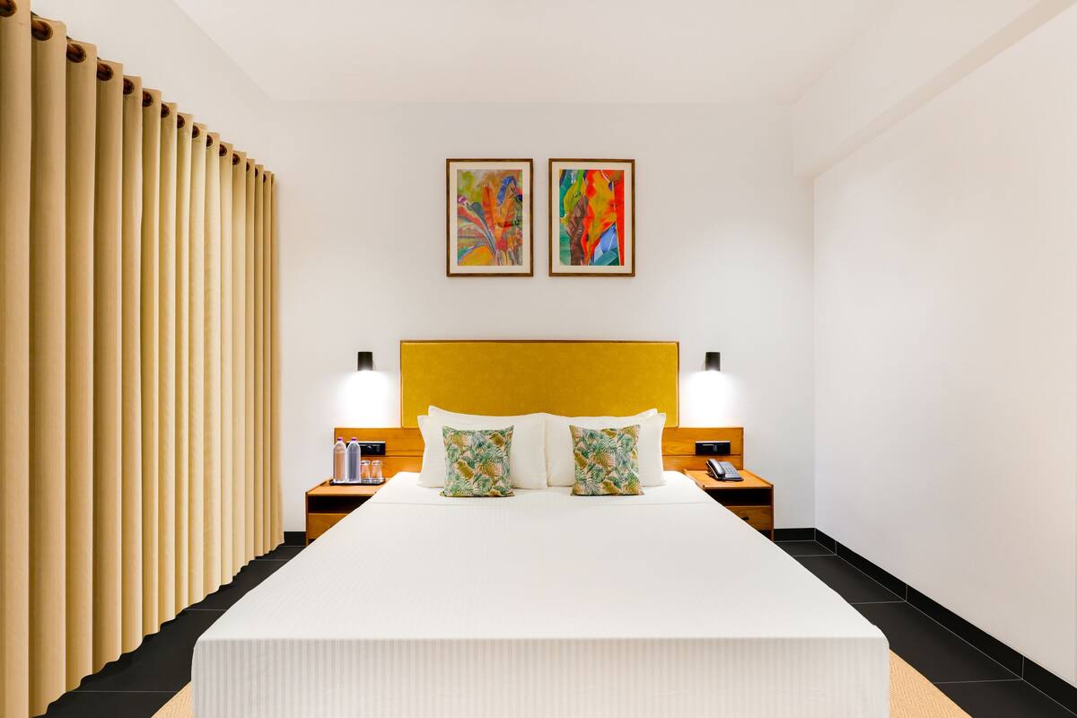 Habitación de hotel con una cama grande, dos mesitas de noche, un cabecero amarillo y dos cuadros abstractos en la pared.