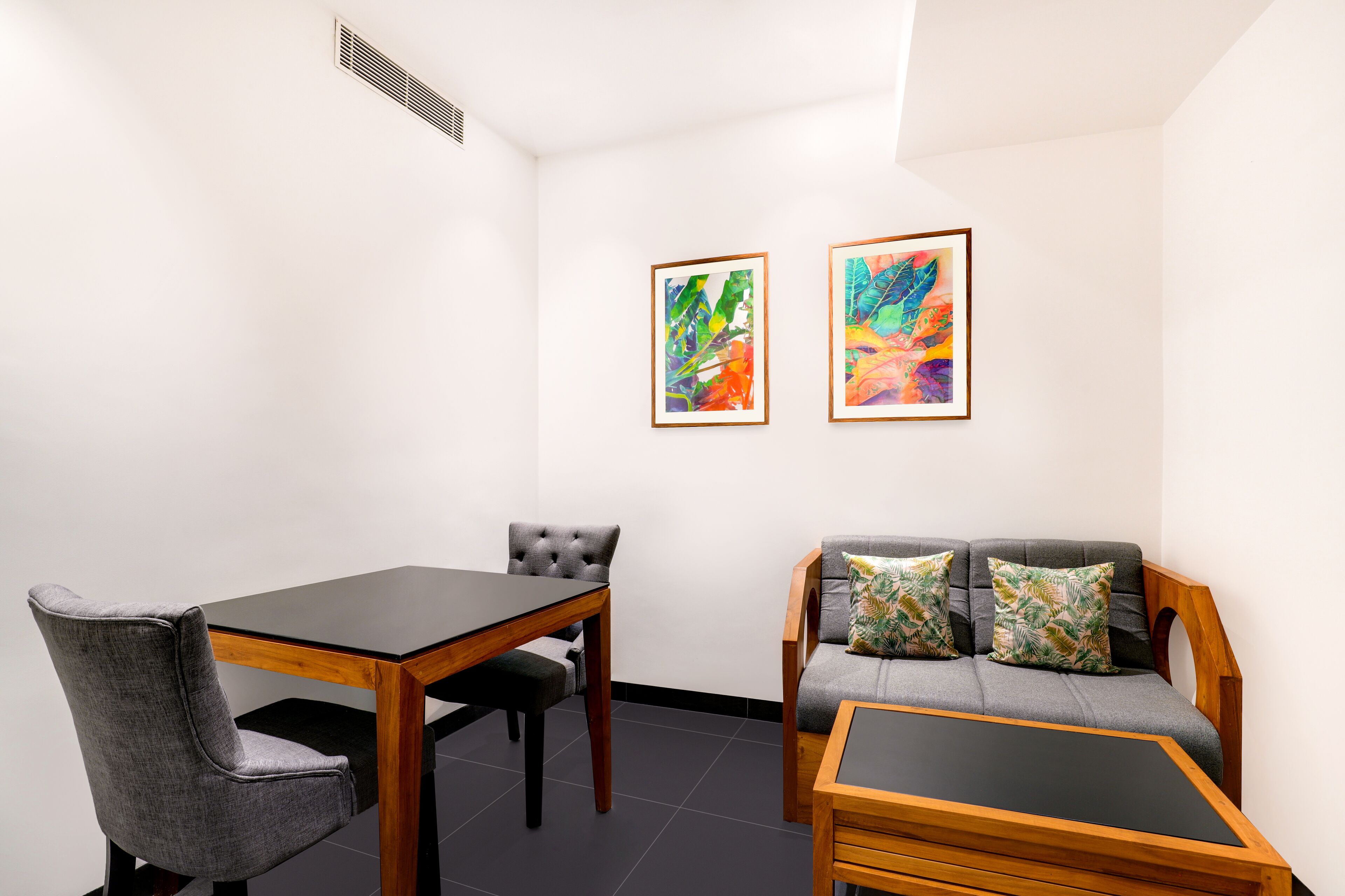 Een kamer met een eettafel, twee stoelen, een bank met kussens, en twee ingelijste abstracte schilderijen aan de muur.