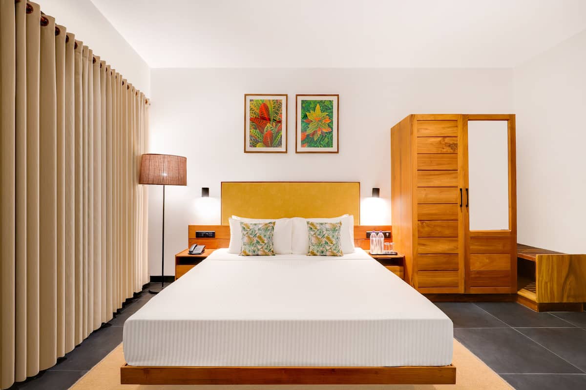 Habitación de hotel con una cama grande, cabecera de madera, dos cuadros colgados en la pared, un armario de madera y una mesita de noche con una lámpara.