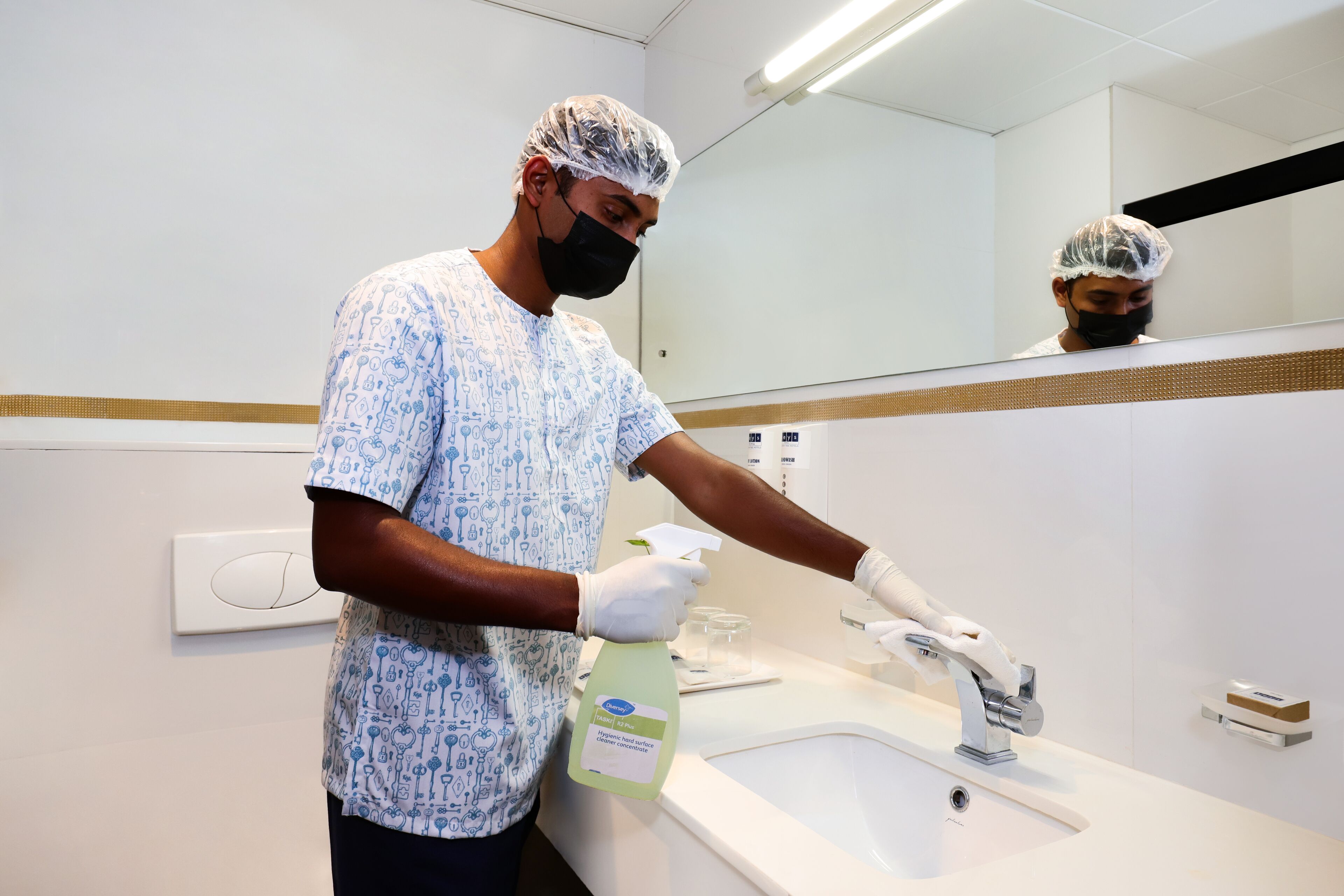 Une personne portant une chemise à motifs et un filet à cheveux, utilisant un vaporisateur dans une salle de bain moderne dotée d’un grand miroir et d’un lavabo blanc.