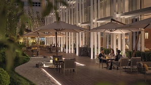 Un patio al aire libre con mesas y sillas bajo sombrillas, rodeado de vegetación y un edificio con grandes ventanales.